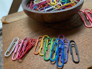 A rainbow-colored array of paper clips on a wooden box w/ a bowl of paper clips in the background).